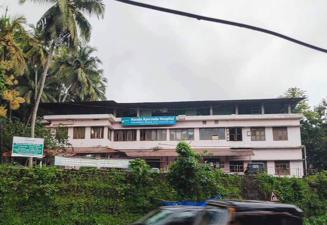 Kerala Ayurveda Hospital building surrounded by greenery and palm trees, with a cloudy sky.