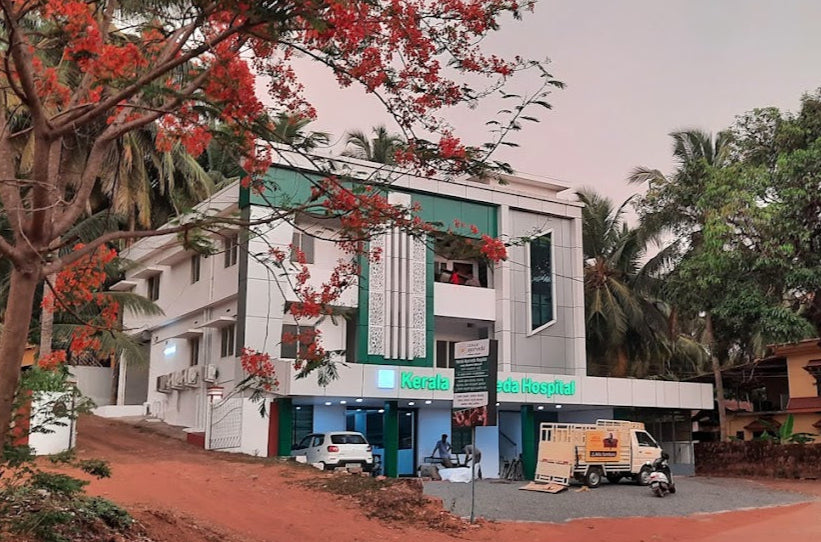 A modern hospital building with green accents surrounded by trees and a parked white vehicle.