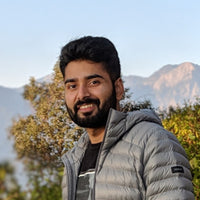 A man with a beard smiles while wearing a gray jacket against a mountain backdrop.