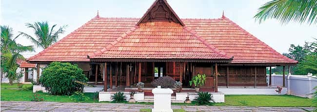 Traditional Kerala house with a sloped tile roof, surrounded by greenery and a manicured lawn.