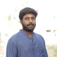 A man with black hair wearing a blue shirt stands outdoors with a blurred background.