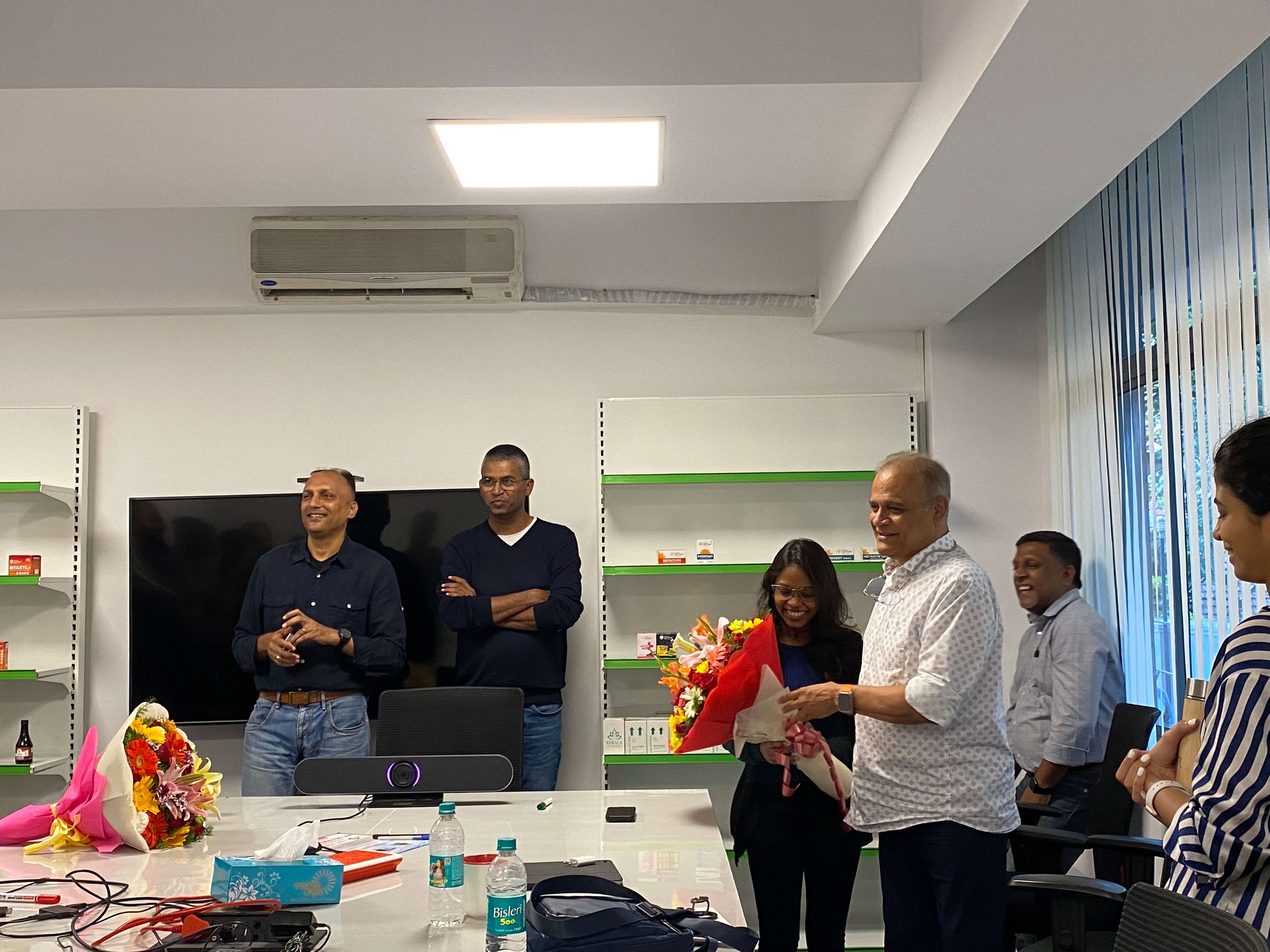 A group of five people in a modern office celebrating with flowers and gifts around a conference table.
