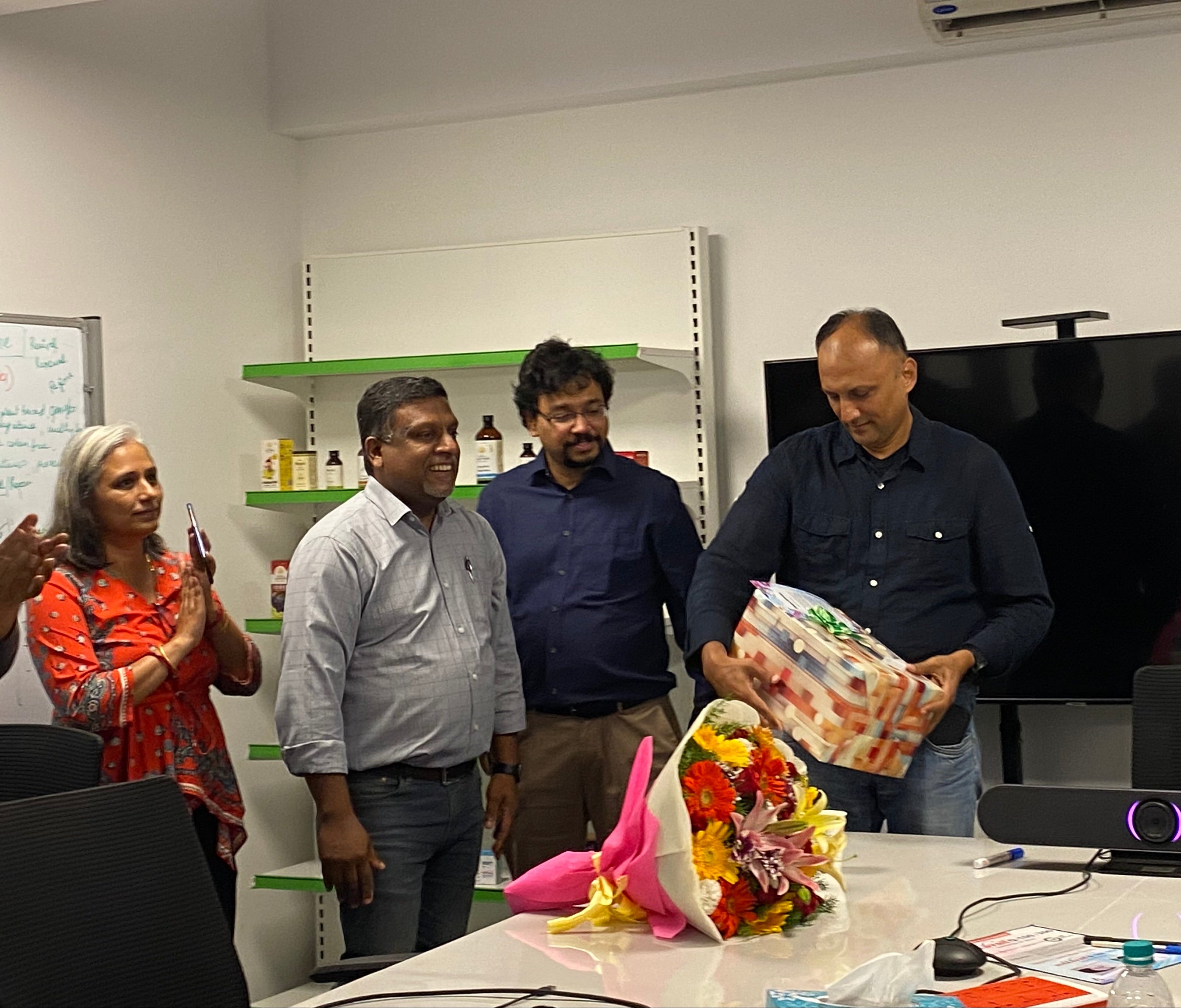 Group of four people in an office setting, with one holding a gift box and flowers on the table.