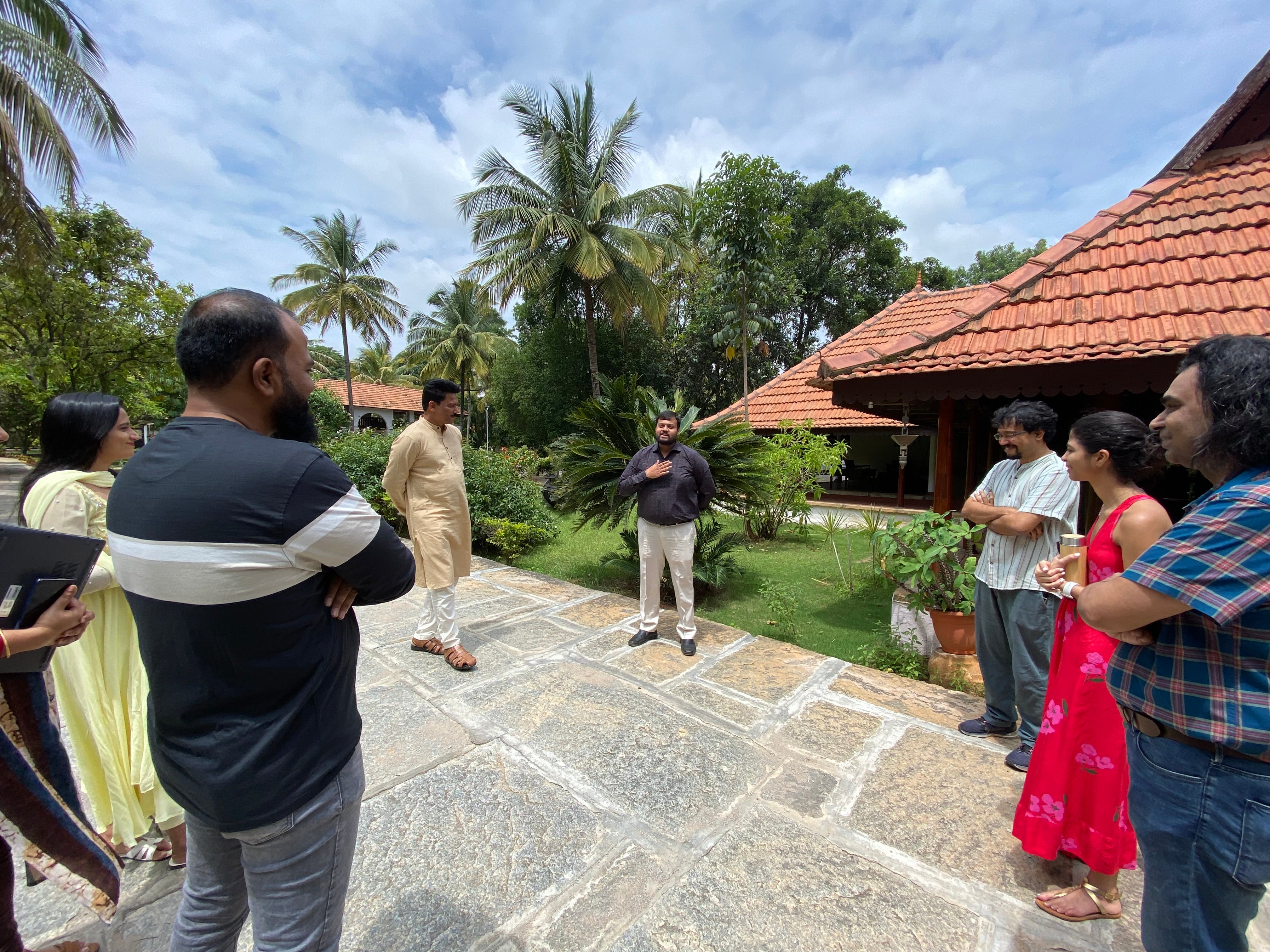 A group of people listening to a speaker in a garden setting with palm trees and traditional architecture.