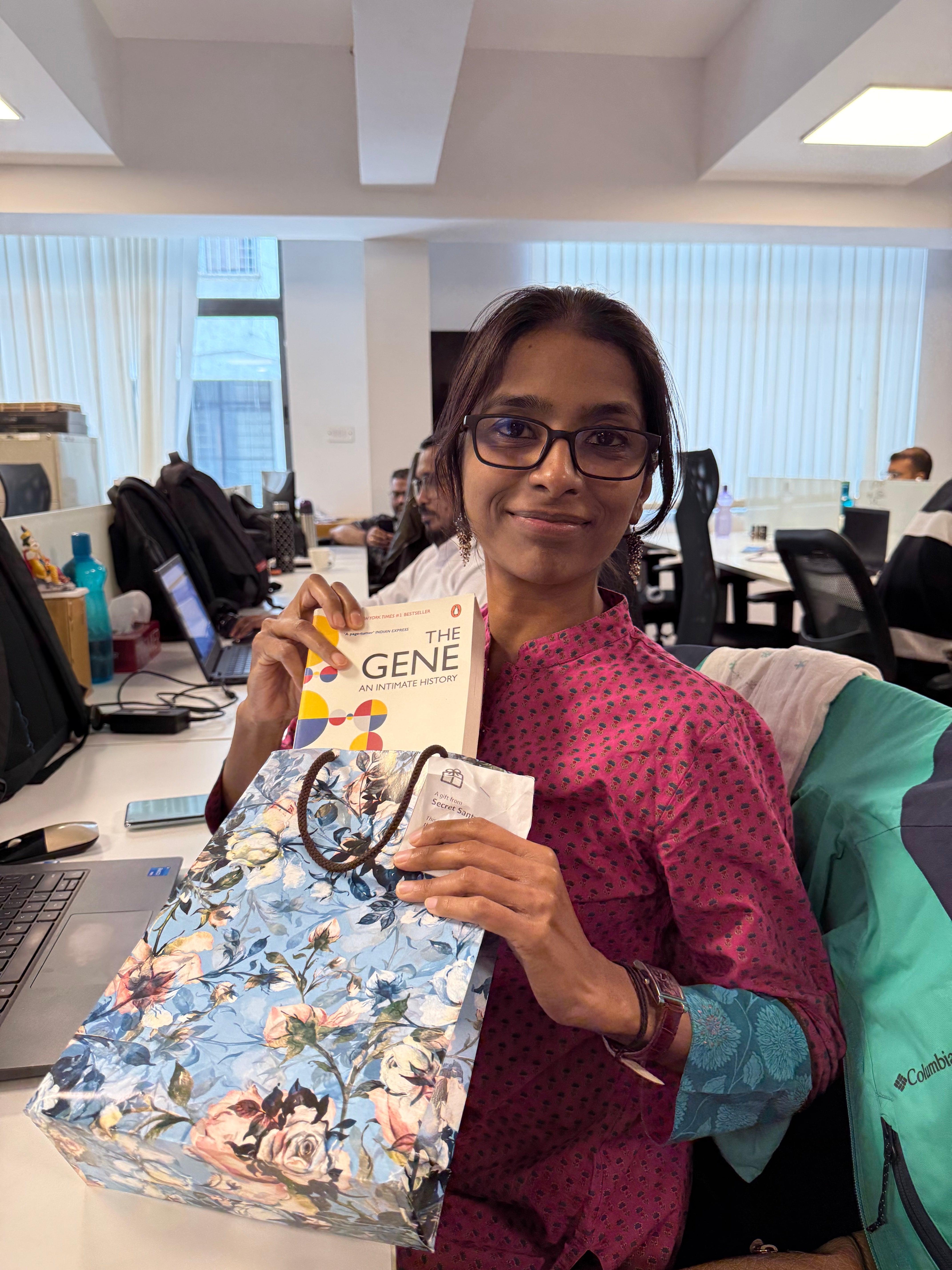 A woman in a pink kurta holds a book titled "The Gene" and a floral gift bag at an office.