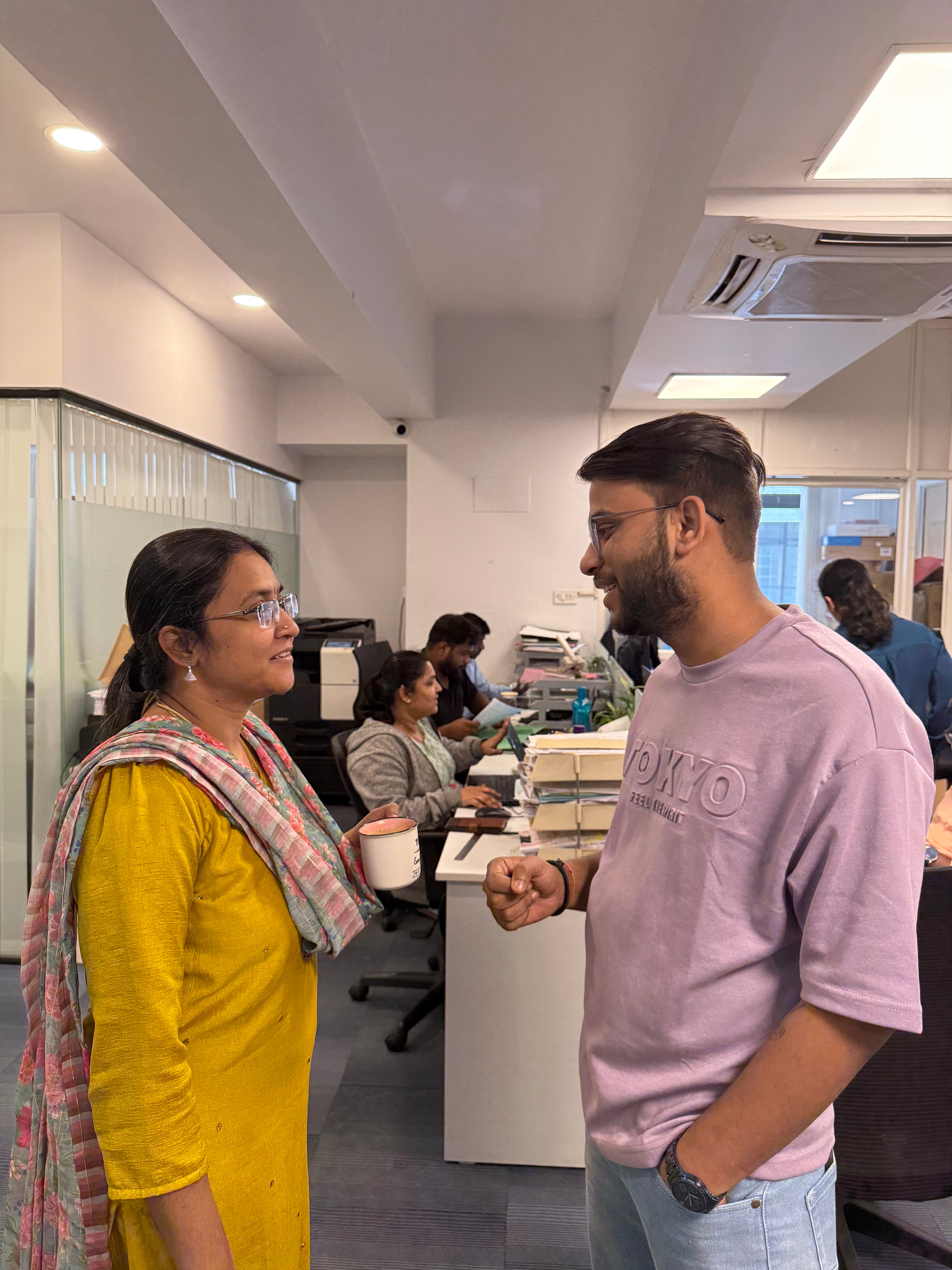 Two people engaged in conversation in an office environment with colleagues working in the background.