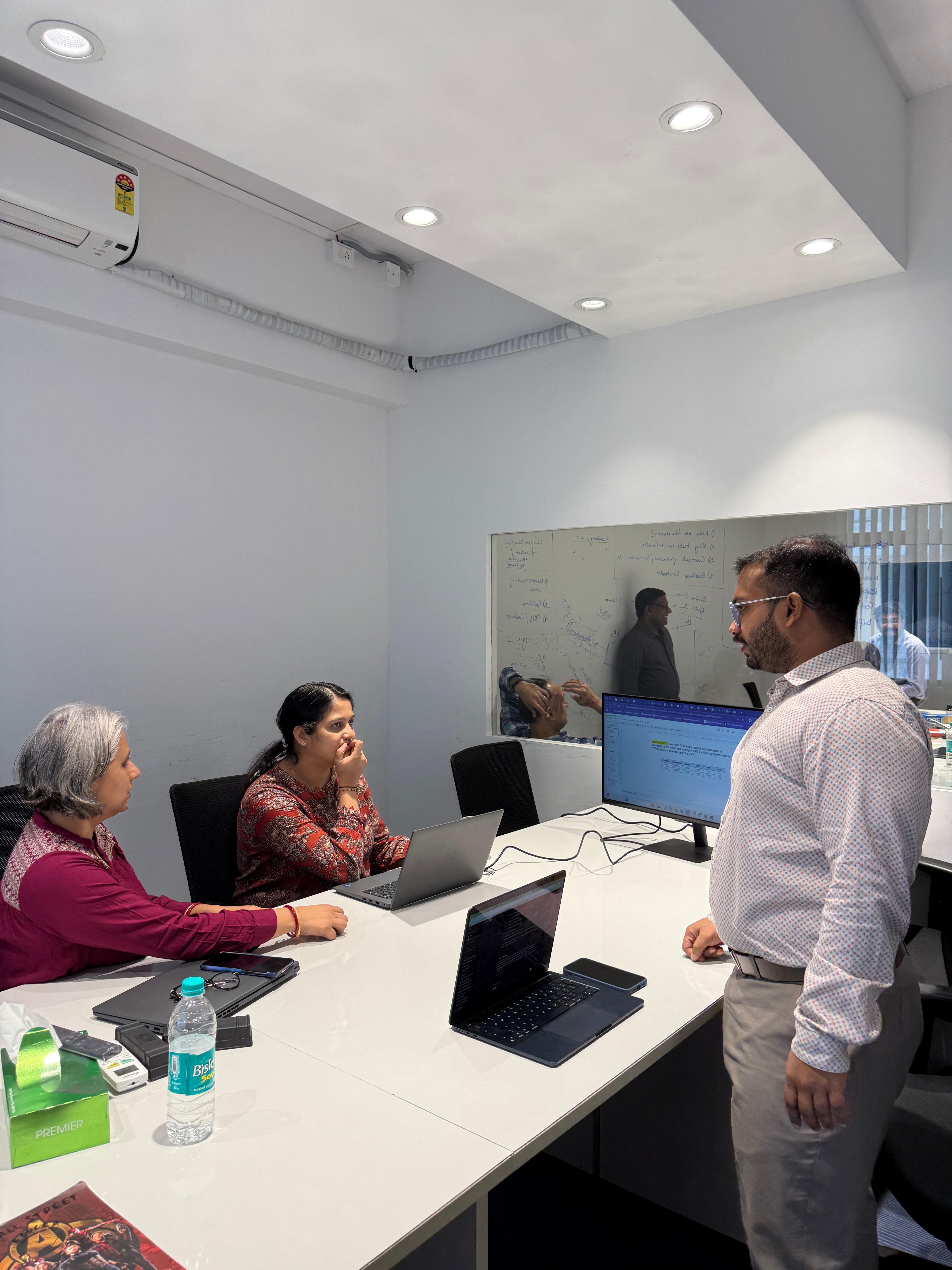 A man stands in a meeting room with two women seated at a table, discussing over laptops.
