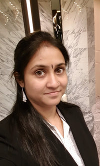 A woman with long black hair in a black blazer, standing in a marble-walled interior.