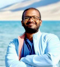 Man with glasses smiling by a body of water, wearing a blue jacket with pink accents.