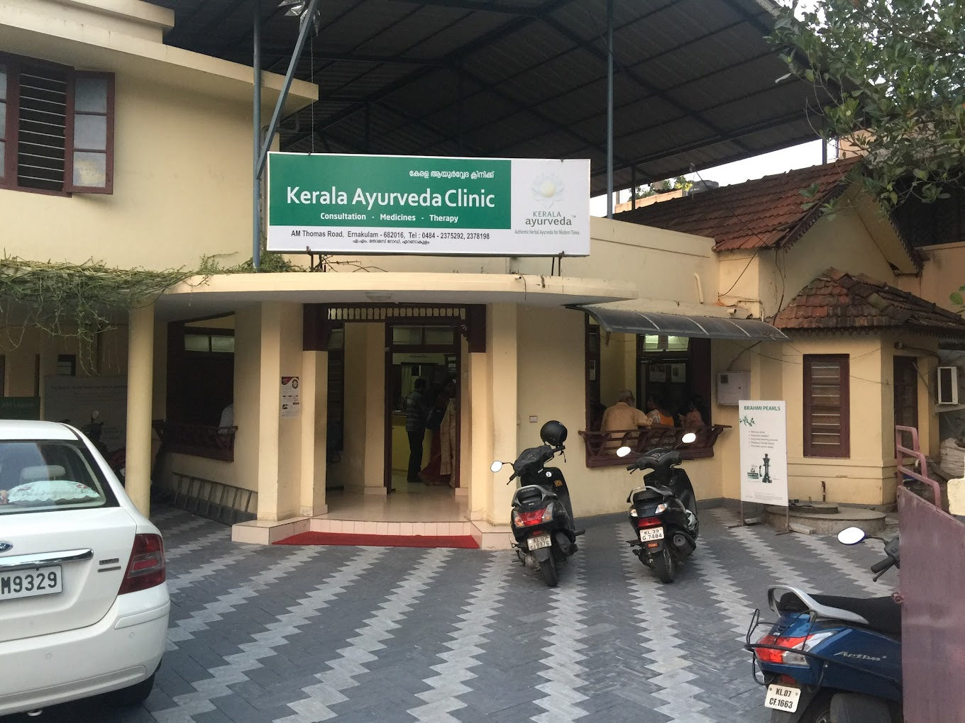 Entrance of Kerala Ayurveda Clinic with two parked motorcycles and a white car in the foreground.