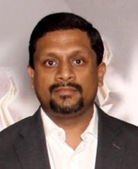 A man in a dark suit and white shirt posing against a soft-focus background.