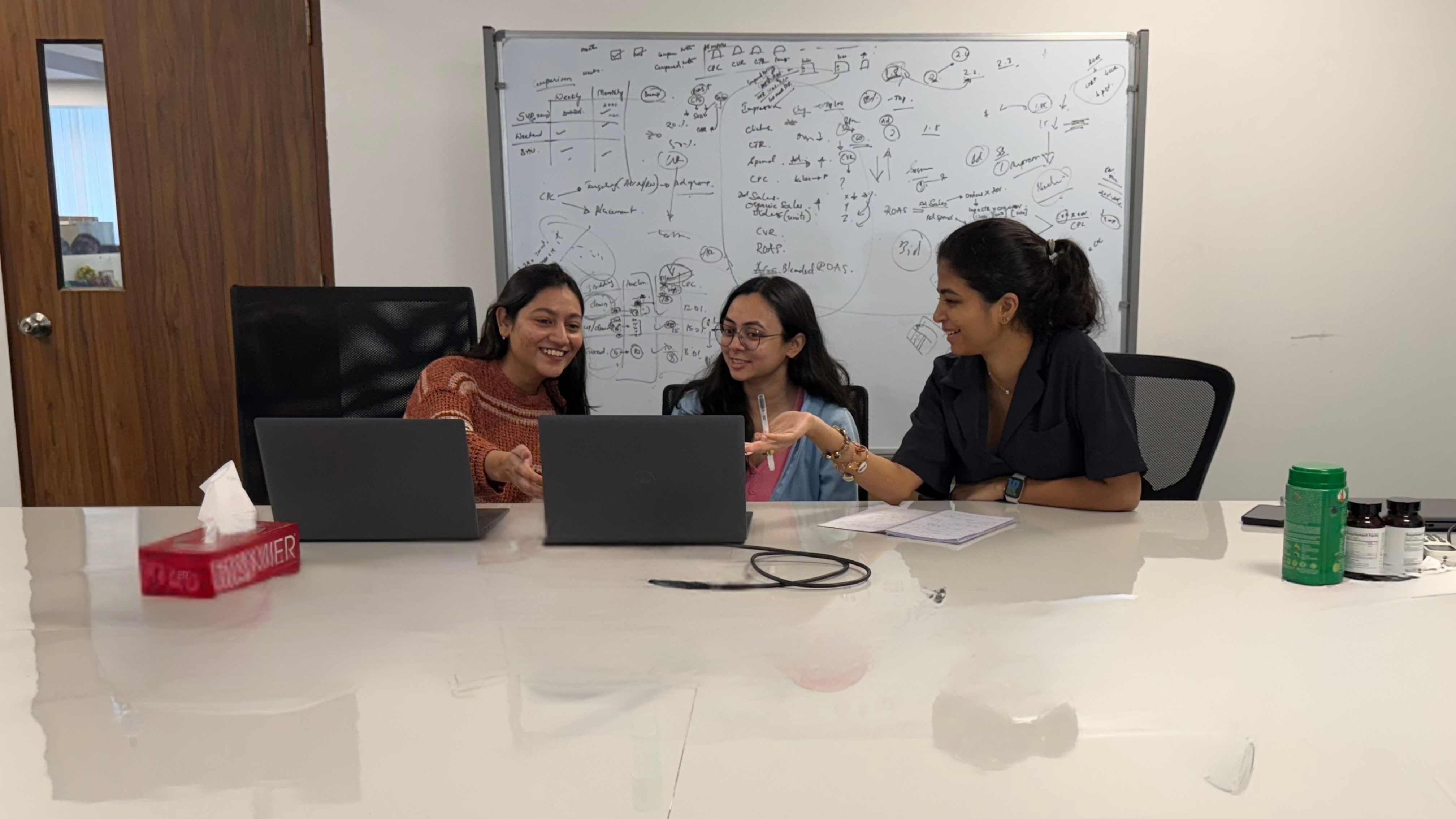 Three women working together at a conference table with laptops and a whiteboard filled with notes in the background.