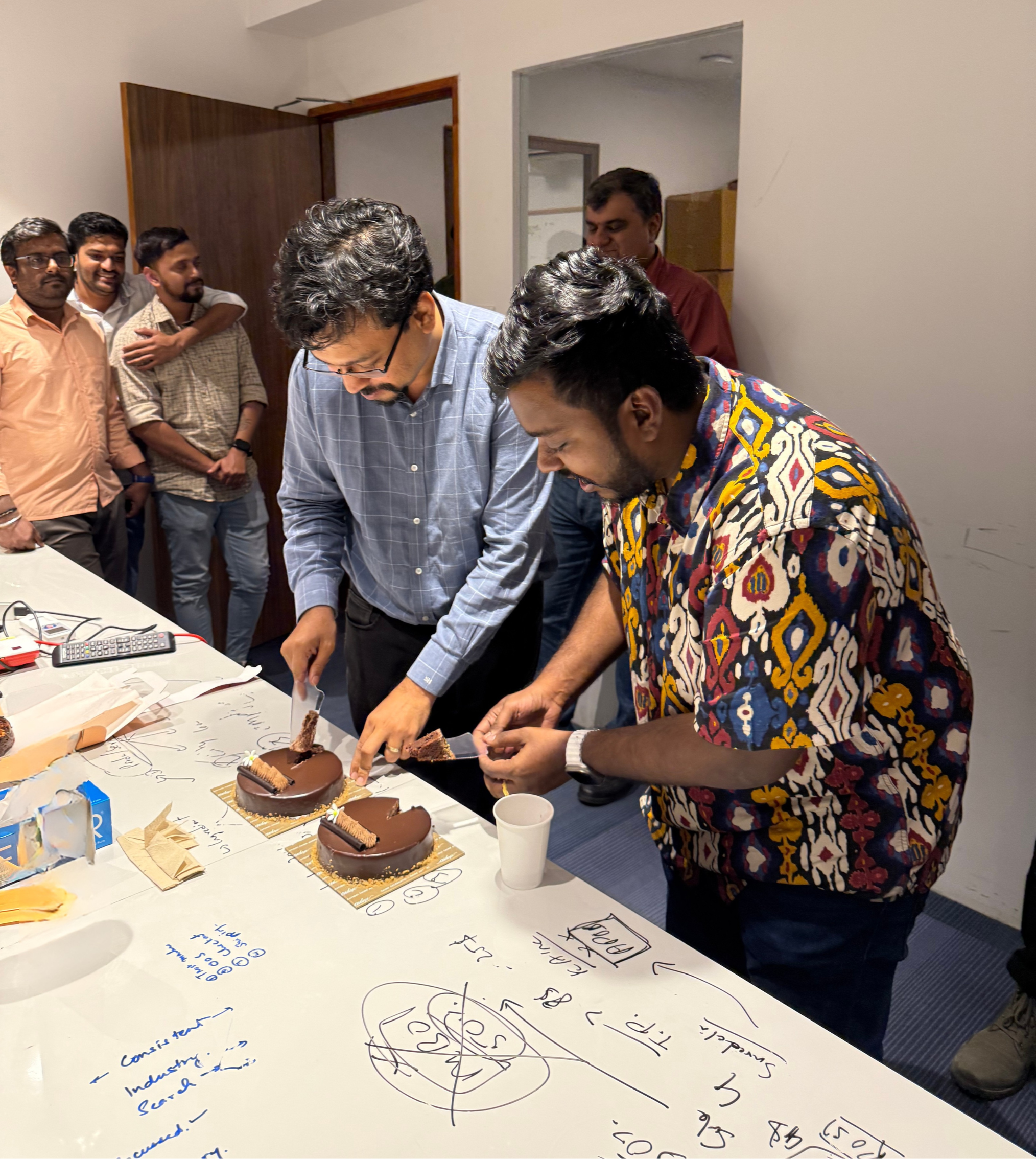 Two men cutting chocolate cakes on a table surrounded by coworkers in an office setting.