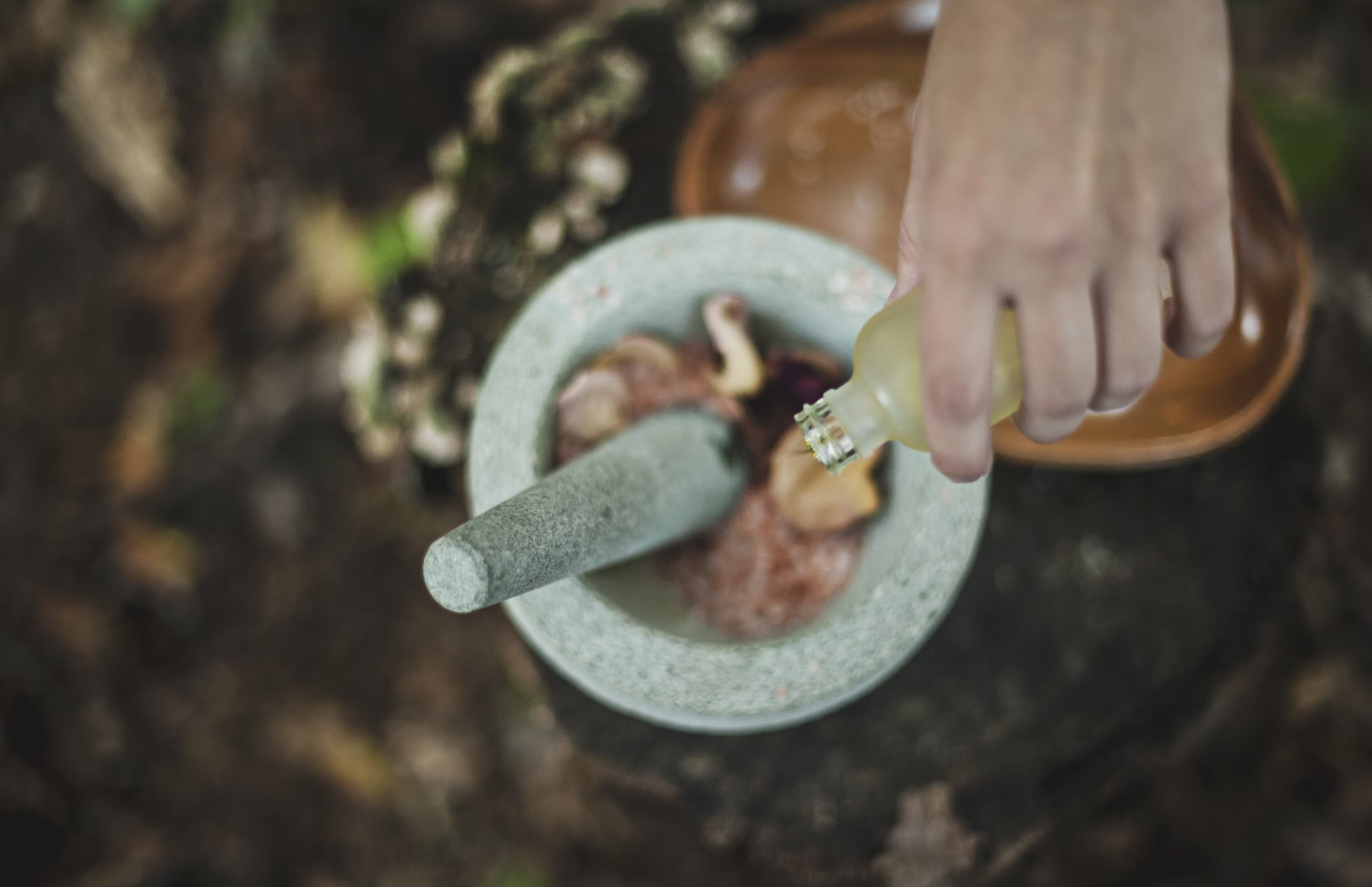 A hand pours oil into a stone mortar containing herbs and spices on a wooden surface.