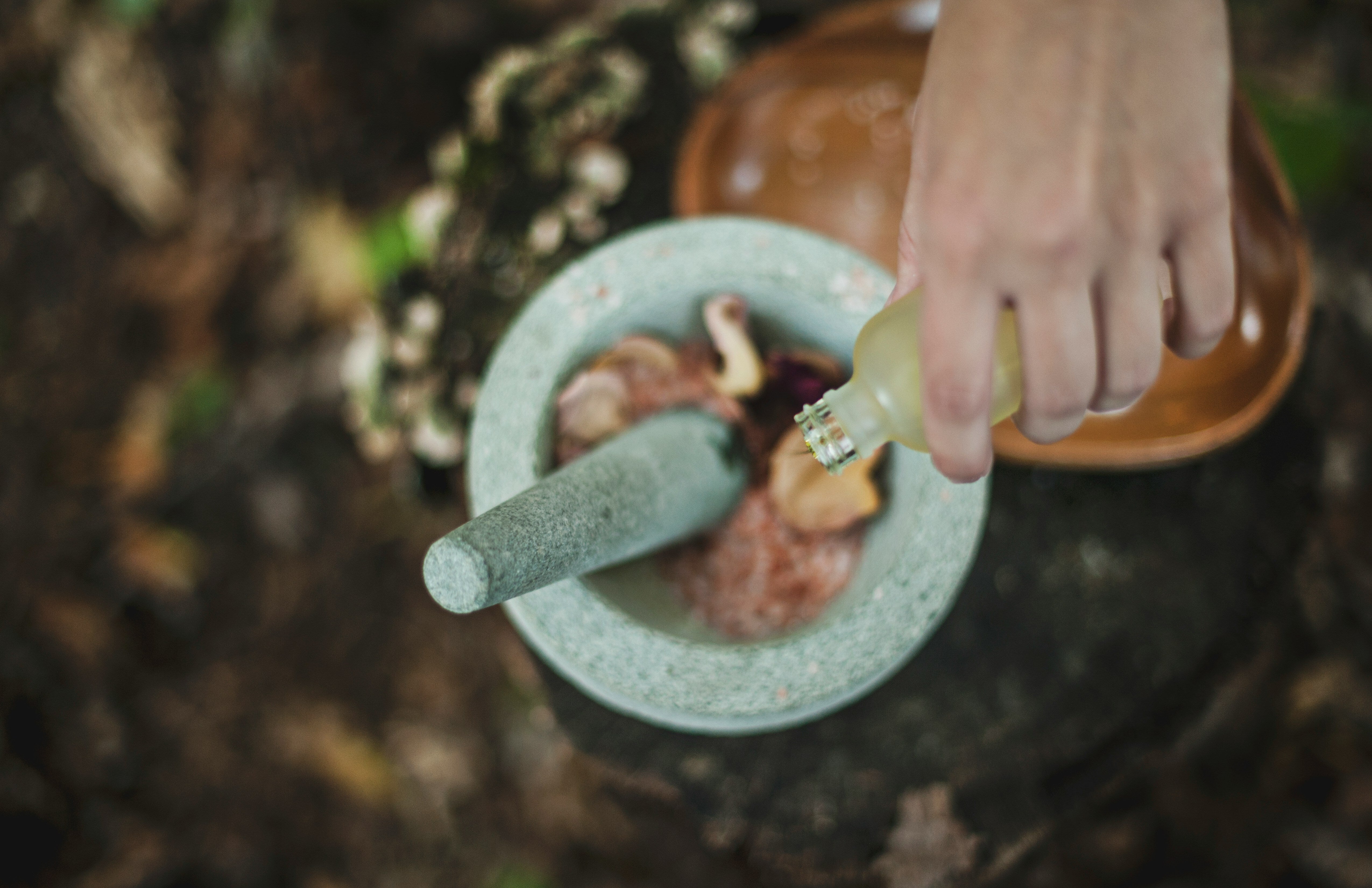 A hand pours liquid into a mortar containing various ingredients on a wooden surface.