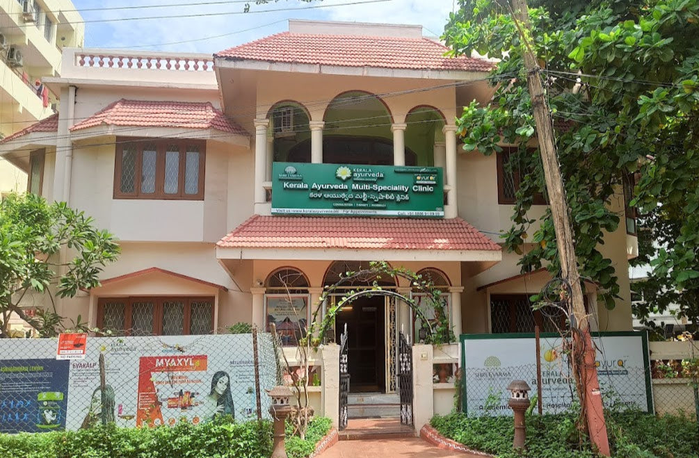 Kerala Ayurveda Multi-Specialty Clinic building with a green banner and lush greenery surrounding it.